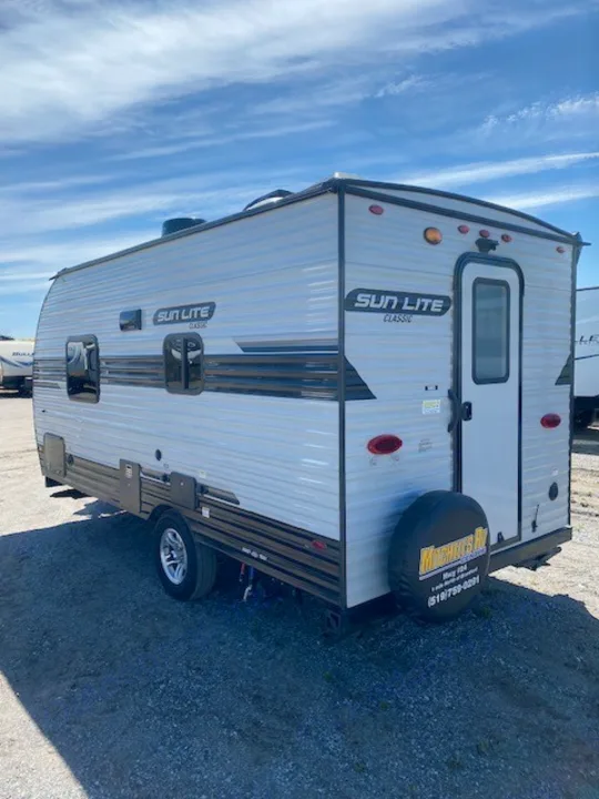 Light gray Sun Lite Classic travel trailer for RV rental. Seen from rear, parked on gravel under a blue sky.. Sunset Park RV Sun Lite 2021