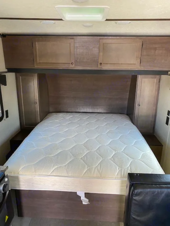 Inside an RV rental: a cozy bedroom with a white quilted queen mattress, overhead cabinets, and side storage.. Sunset Park RV Sun Lite 2021