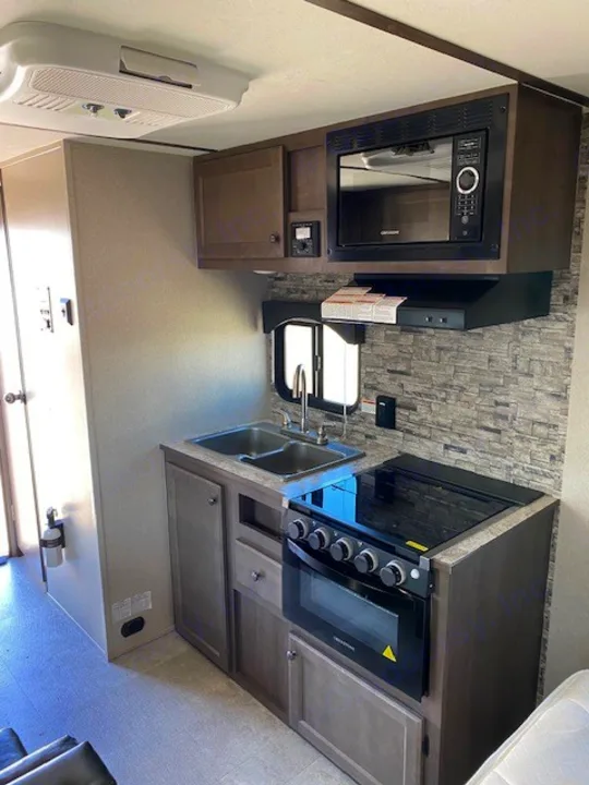 RV rental kitchen with stove, oven, microwave, double sink, cabinets, stone backsplash, and ceiling AC unit.. Sunset Park RV Sun Lite 2021