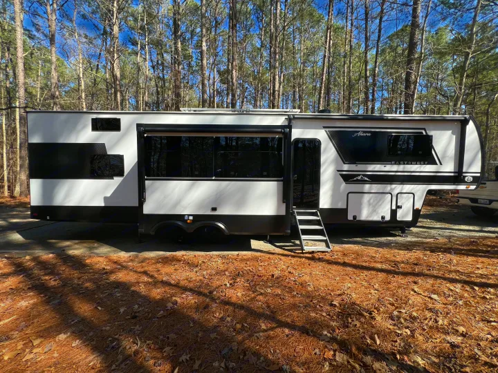 Modern black & white fifth-wheel RV rental parked in a sunlit forest campsite. Perfect for nature adventures.. Forest River East to West Ahara 390DS 2025