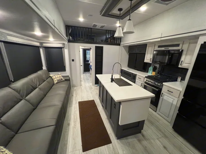 Modern RV rental interior: large grey sofa, white kitchen island with sink, oven, fridge, and overhead loft. Bright and inviting.. Forest River East to West Ahara 390DS 2025