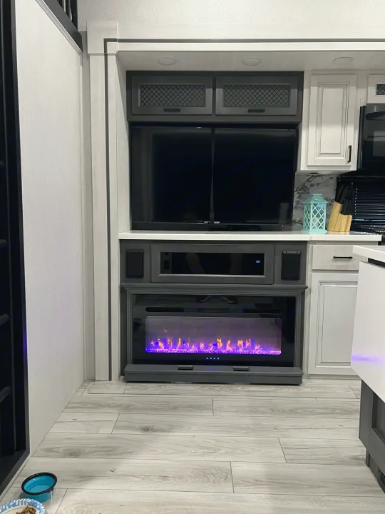 RV rental interior featuring a TV, electric fireplace with vibrant purple flames, kitchen cabinets, and light wood floors.. Forest River East to West Ahara 390DS 2025