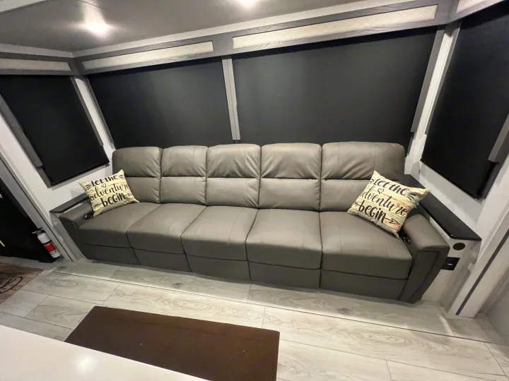 Cozy RV rental living space with a large gray reclining sofa, wood-look floor, and blackout shades.. Forest River East to West Ahara 390DS 2025