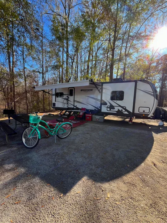 RV rental campsite: A white travel trailer with awning, picnic table, green bike, and red chairs under sunny trees.. Sporttrek Touring Edition 2024