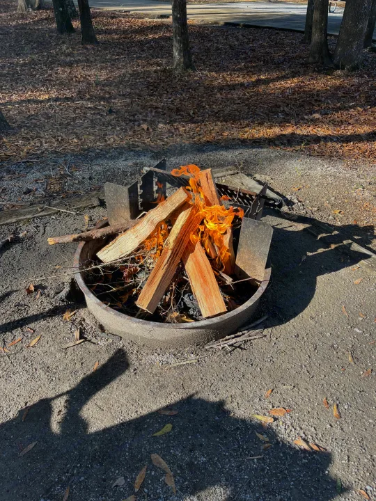 Daytime campfire in a metal pit at a gravel RV rental campsite, surrounded by trees and leaves.. Sporttrek Touring Edition 2024
