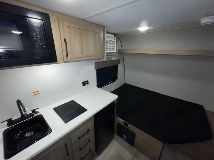 Compact RV rental interior featuring a modern kitchen with microwave, sink, cooktop, and a cozy bed area.. Keystone RV Coleman 13B 2025