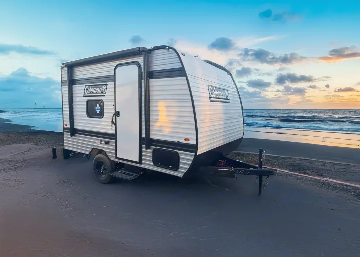 A Coleman travel trailer, ideal for RV rental camping, sits on a sandy beach at sunset. Perfect for outdoor adventures.. Keystone RV Coleman 13B 2025