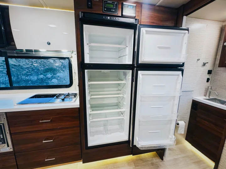 Modern and spacious rv rental kitchen interior with an open, empty fridge, cooktop, sink, and wood cabinetry.. Winnebago Navion 2022