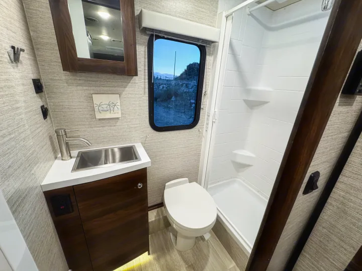 This compact, modern RV rental bathroom features a bright shower, toilet, and sink with a window offering nature views.. Winnebago Navion 2022
