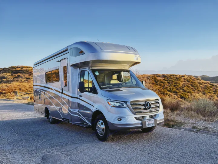 This modern silver Mercedes-Benz Sprinter Class C RV rental is parked on a scenic road amidst dry hills under a clear sky.. Winnebago Navion 2022