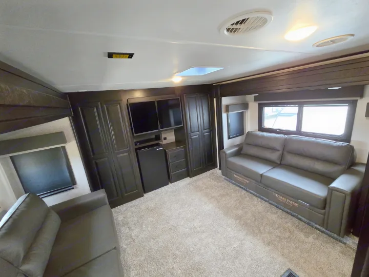 RV rental living area with dark wood cabinets, dual gray Thomas Payne sofas, TV, minifridge, skylight, and plush carpet.. Open Range Highland Ridge 2021