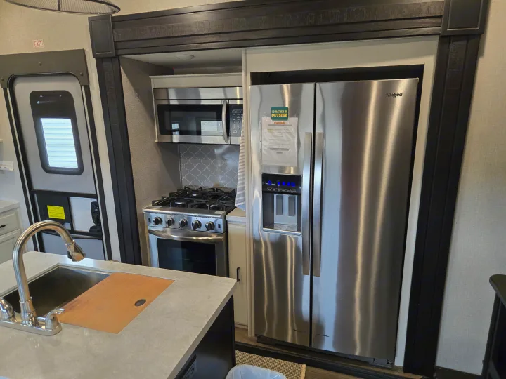 Modern RV rental kitchen featuring a large stainless steel fridge, microwave, oven, and sink with a cutting board.. Open Range Highland Ridge 2021
