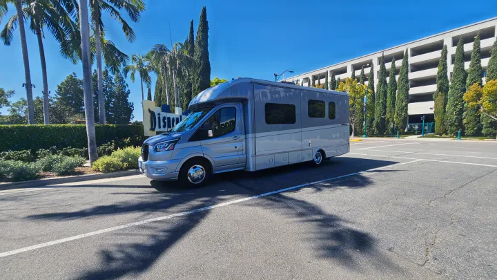 A gray **rv rental** is parked at the iconic Disneyland entrance sign. Sunny day with palm trees & a clear blue sky.. Tiffin Motorhomes Allegro Midas 24MT 2024