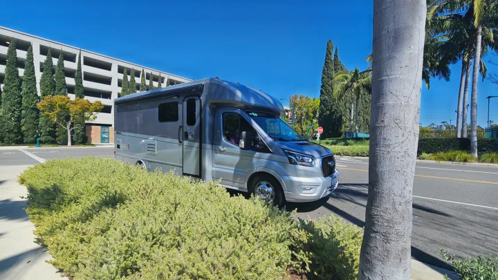 Modern silver RV rental showcased on a sunny day, parked curbside with lush greenery and a parking garage.. Tiffin Motorhomes Allegro Midas 24MT 2024