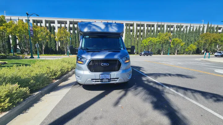 Silver Tiffin Ford Transit RV rental parked on a sunny road, with a large building and cypress trees in the background.. Tiffin Motorhomes Allegro Midas 24MT 2024