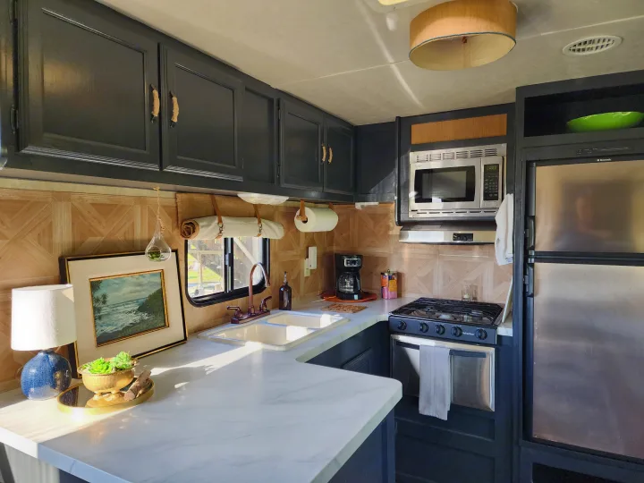 Explore this stylish RV rental kitchen. Features dark cabinetry, white marble-look counters, sink, stovetop, microwave, and fridge.. Forest River Wildwood 2017
