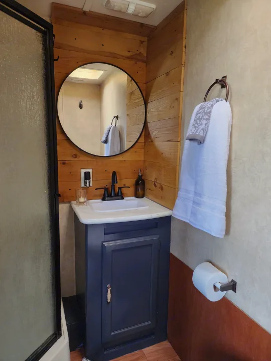 Renovated RV rental bathroom with stylish vanity, sink, round mirror, shower, and rustic wood paneling.. Forest River Wildwood 2017