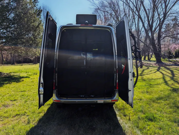 Rear view of a silver RV rental van with wide open doors revealing its interior and a bike rack, parked on green grass.. Thor Sanctuary 2021