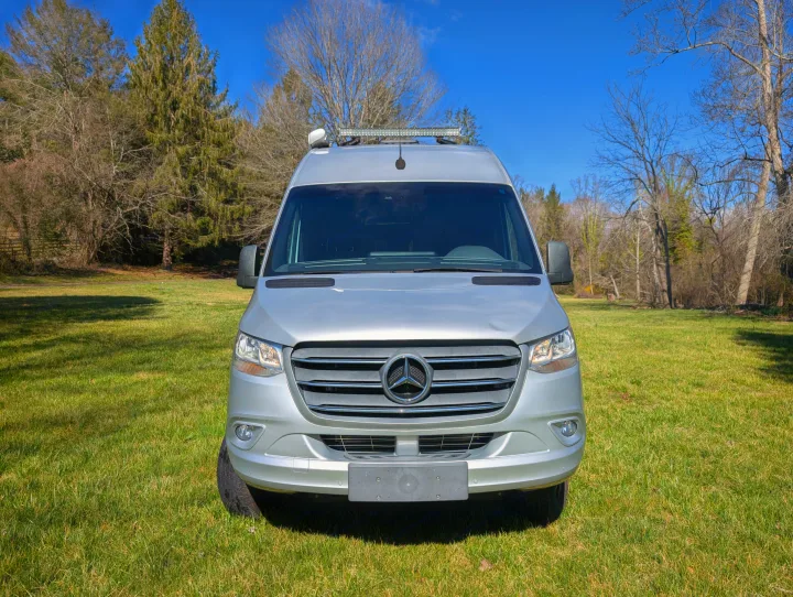 Front view of a silver Mercedes Sprinter van, perfect for **rv rental**, parked in a sunny grassy field.. Thor Sanctuary 2021