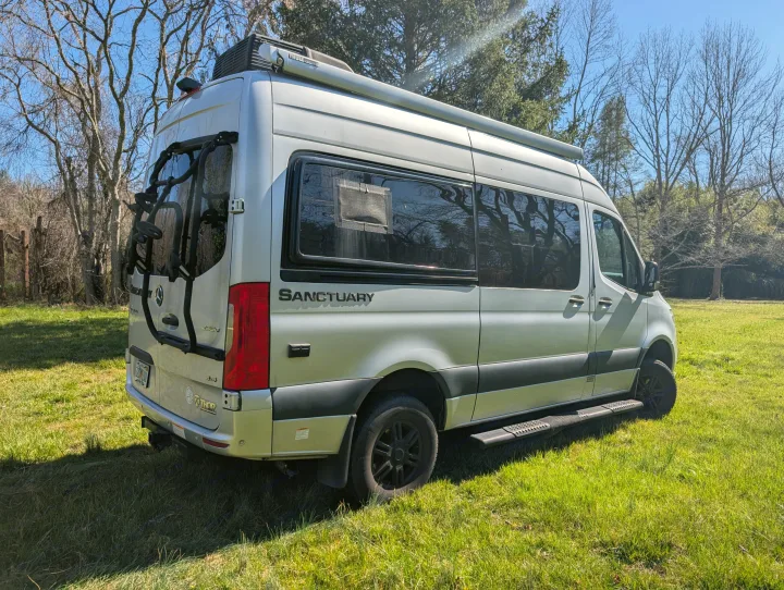 Silver Sanctuary RV rental, a sleek camper van with bike rack and roof awning, parked in a sunny grassy field.. Thor Sanctuary 2021