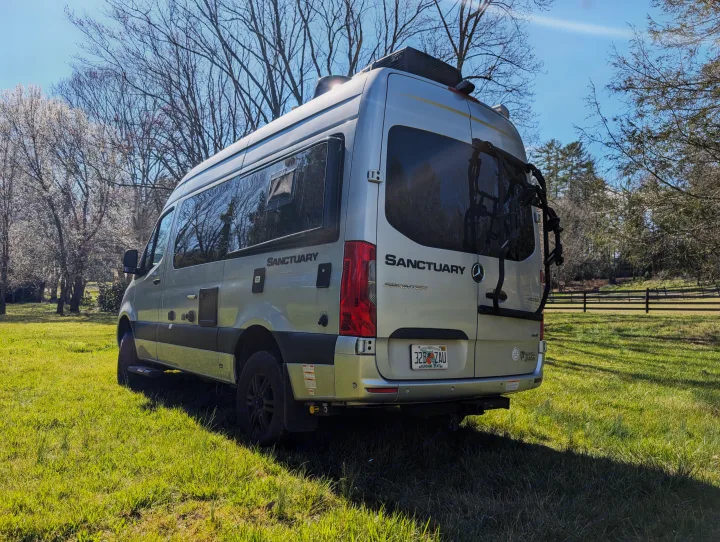 Silver Sanctuary RV rental, a Mercedes-Benz Sprinter camper van, parked in a sunny grassy field with trees.. Thor Sanctuary 2021