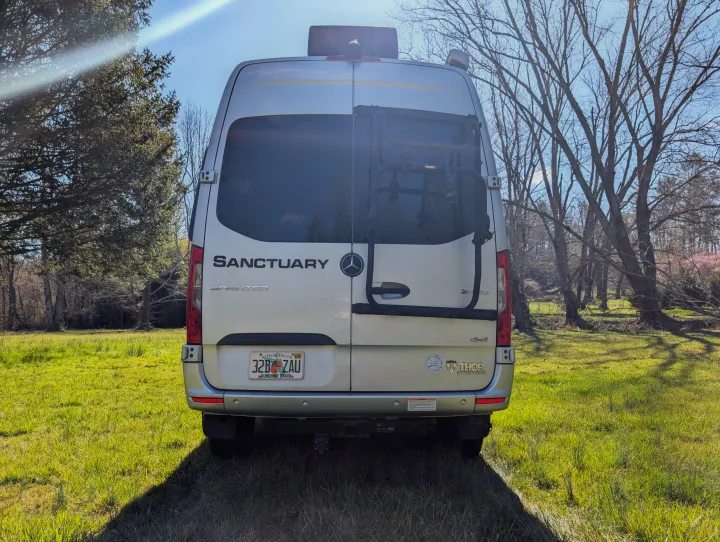 Rear of a silver Thor Sanctuary Sprinter RV rental with a bike rack, parked in a sunny, grassy field.. Thor Sanctuary 2021