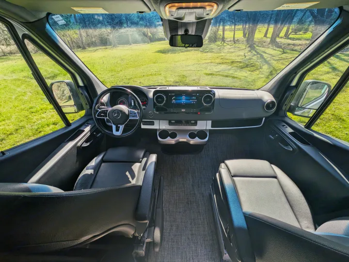 Spacious RV rental's front cabin interior with dashboard, seating, and a sunny green landscape view through the windshield.. Thor Sanctuary 2021