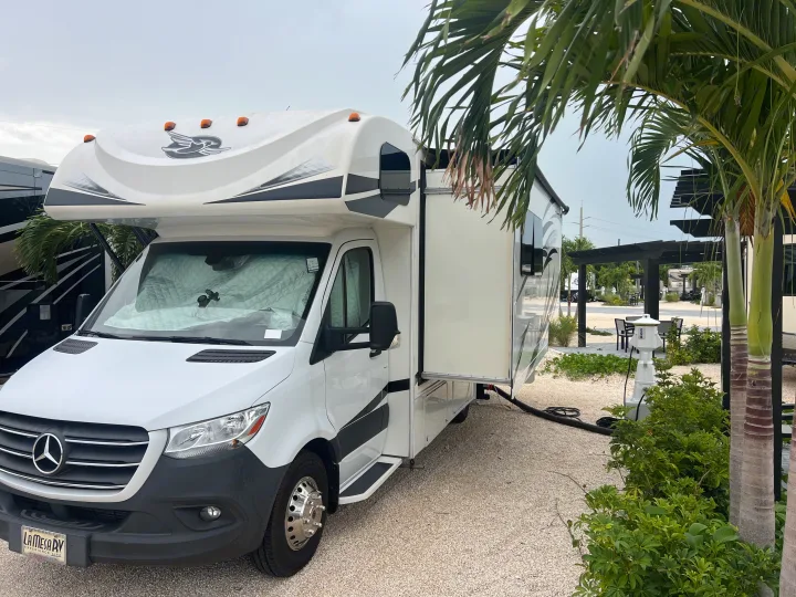 White Mercedes-Benz Class C RV rental at an RV park. Slide-out extended, on a gravel site with palm trees.. Jayco Melbourne 2020