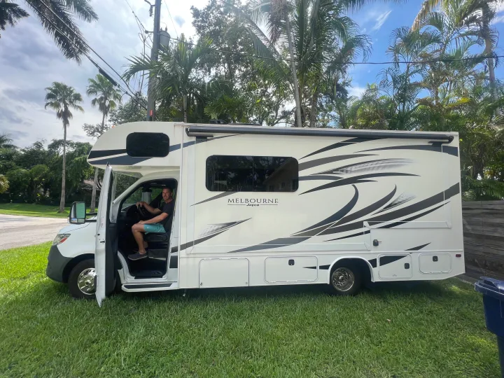 White Melbourne Jayco rv rental motorhome parked on grass. Man in driver's seat. Palm trees line the road.. Jayco Melbourne 2020