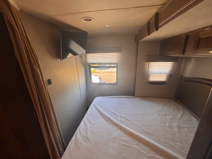 RV rental bedroom featuring a comfortable bed, wall-mounted TV, windows with blinds, and practical overhead storage.. Jayco Melbourne 2020