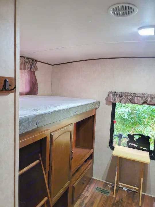 RV rental interior featuring a bunk bed with a white mattress, wooden storage unit, ladder, and a window with green outdoor view.. Forest River Wildwood Heritage Glen Lite 2014