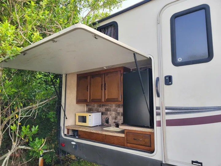 RV rental with an outdoor kitchen setup featuring a microwave, sink, fridge, and cabinets, surrounded by lush trees.. Forest River Wildwood Heritage Glen Lite 2014