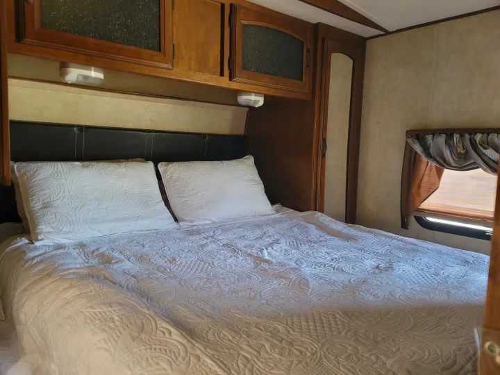 RV rental bedroom with a white quilted bed, black headboard, overhead wooden cabinets, and a window.. Forest River Wildwood Heritage Glen Lite 2014