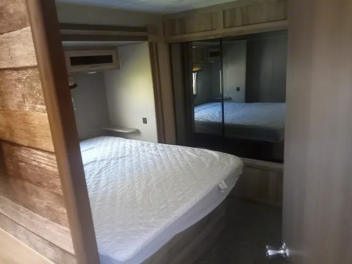 RV rental bedroom featuring a bed with white quilted cover, mirrored closet, and overhead cabinets for storage.. Trail Runner heartland 25sle 2016