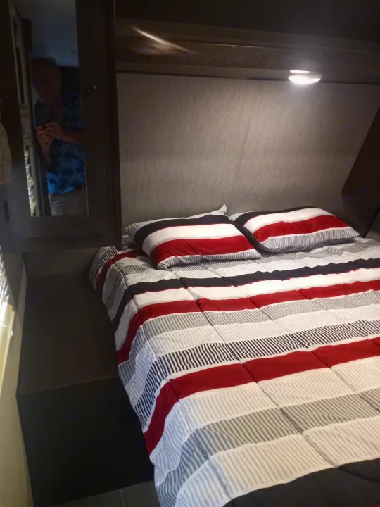 Inside an **rv rental** bedroom: a comfy bed with vibrant red, white, & black striped bedding. Mirror shows photographer.. Trail Runner heartland 25sle 2016