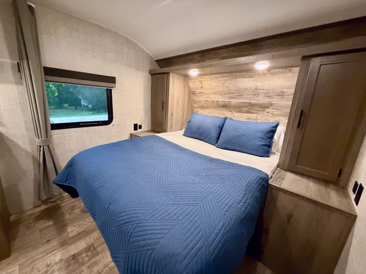 Inside an RV rental: a modern bedroom with a blue quilted bed, wood accents, overhead cabinets, and a window view.. Gulf Stream Kingsport 2021