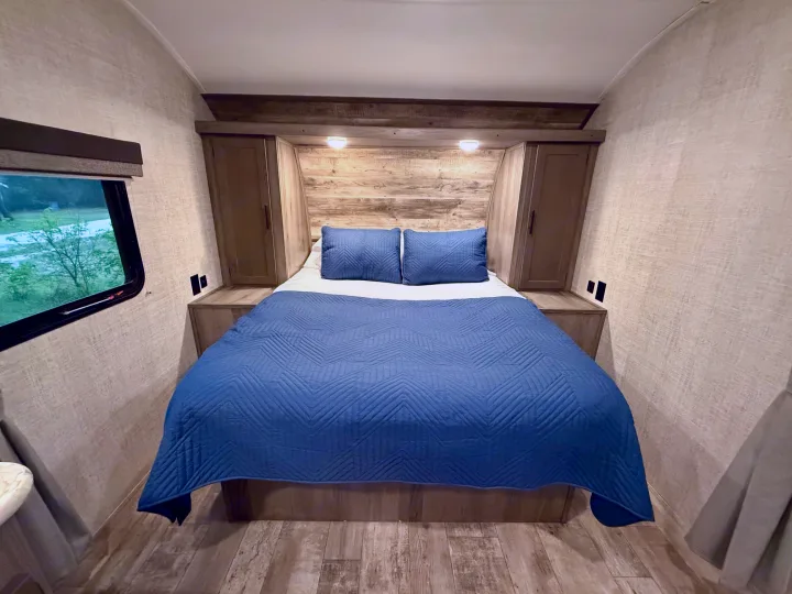 Inside a cozy RV rental bedroom: bed with blue quilt, rustic wood headboard, built-in cabinets, overhead lights, and a window.. Gulf Stream Kingsport 2021
