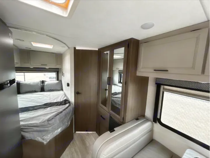 RV rental interior showing a comfortable bed with gray bedding, mirrored wardrobe, overhead cabinets, and a seating area.. GMC/THOR MAGNITUDE 2025