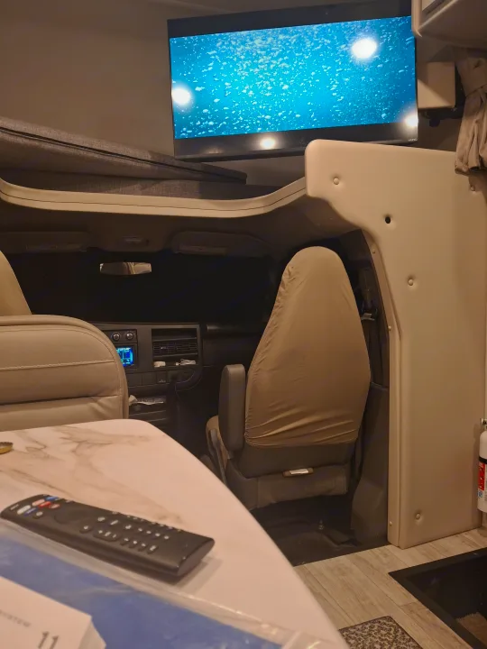 RV rental living space: TV displaying a vibrant blue scene with bubbles, driver's cab, and a table with remote. Ideal for travel.. GMC/THOR MAGNITUDE 2025