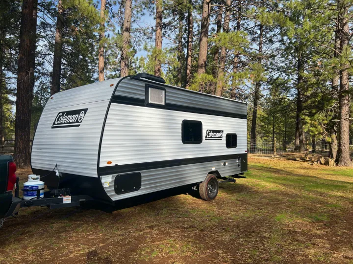 Coleman travel trailer parked amidst pine trees, ideal for an RV rental or camping getaway in nature.. Coleman 17B 2025