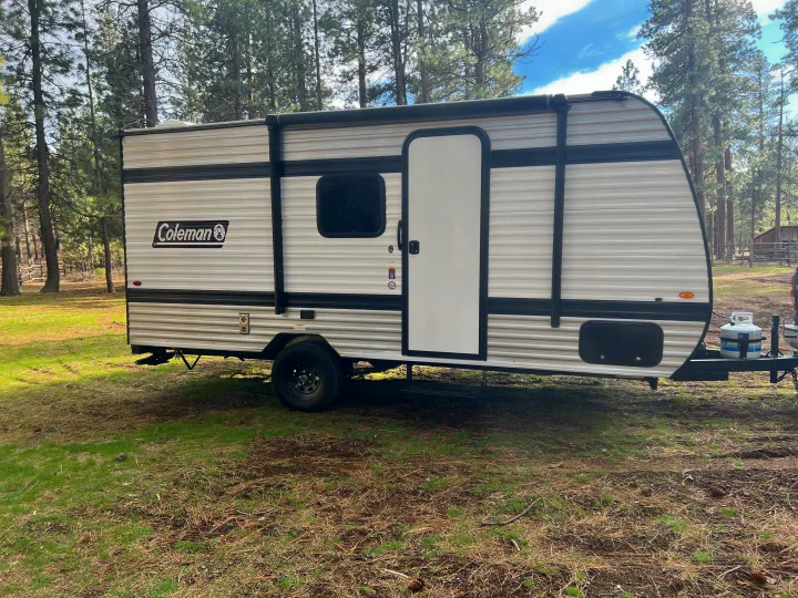 This Coleman travel trailer is an RV rental, parked in a serene, wooded campsite, ready for your next outdoor adventure.. Coleman 17B 2025