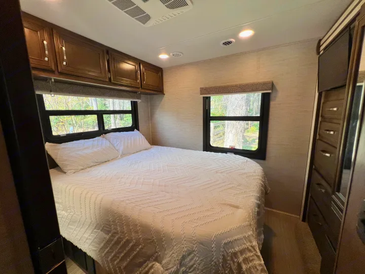 Inviting RV rental bedroom with a white bed, dark wood cabinets, windows showing trees, and built-in storage.. Entegra Coach Esteem 2021