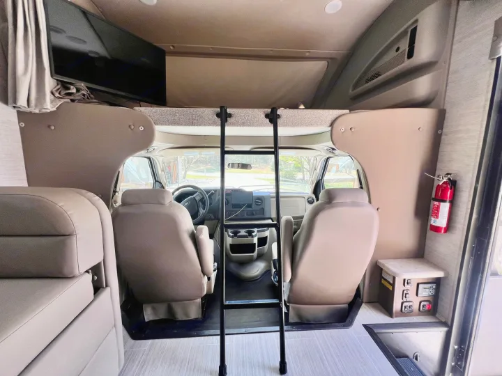 Inside an RV rental: view of the front cockpit, ladder to the over-cab bunk, TV, controls, and fire extinguisher.. Entegra Coach Esteem 2021