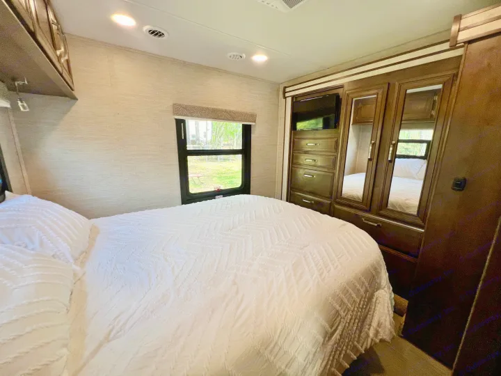 Spacious RV rental bedroom with a bed, white textured bedding, an outdoor window view, and dark wood built-in storage.. Entegra Coach Esteem 2021