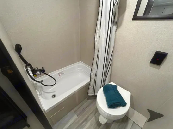 Compact RV rental bathroom featuring a small tub/shower combo, toilet with teal towel, and striped shower curtain.. Other Other 2025