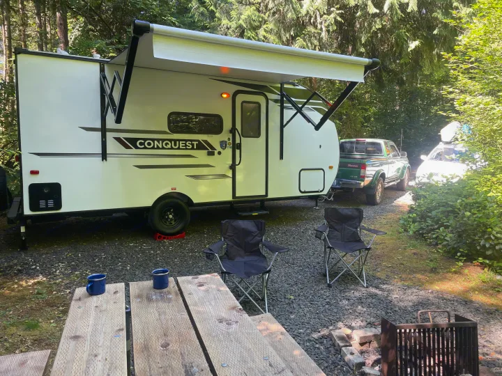 Scenic rv rental setup: a white travel trailer with an extended awning, chairs, and picnic table at a shaded forest campsite.. Gulf Stream Ameri-Lite 2024