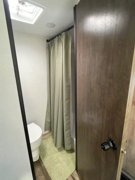 Compact RV rental bathroom with toilet, shower curtain, and wood door. Ideal for your next camper adventure.. Gulf Stream Ameri-Lite 2024