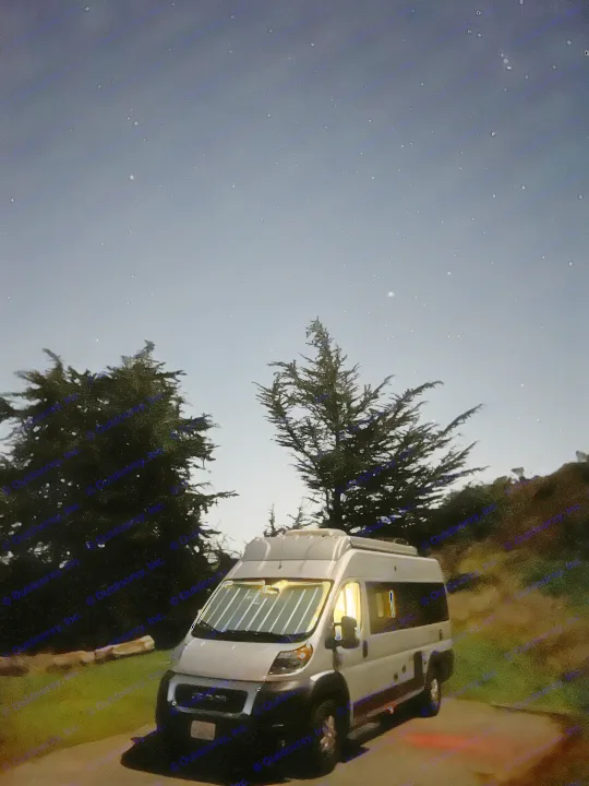 A white camper van, likely an RV rental, illuminates the dark, serene night, parked under a sky full of bright stars, flanked by dark trees.. Thor Motor Coach Sequence 2022