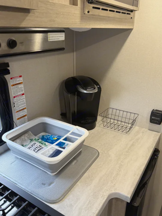 Compact RV rental kitchen. Features a Keurig, collapsible dish drying rack, and crucial safety warning labels.. Forest River Coachmen Catalina Summit 2024