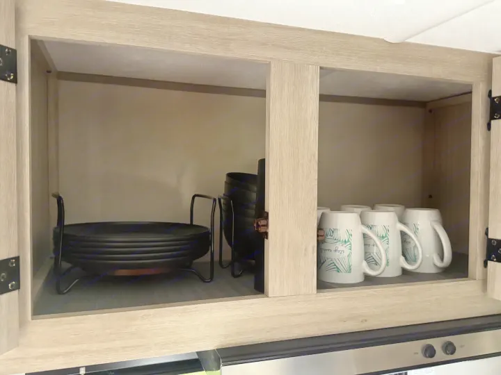 Inside an RV rental kitchen cabinet, showing neatly organized black plates, bowls, and patterned white mugs.. Forest River Coachmen Catalina Summit 2024